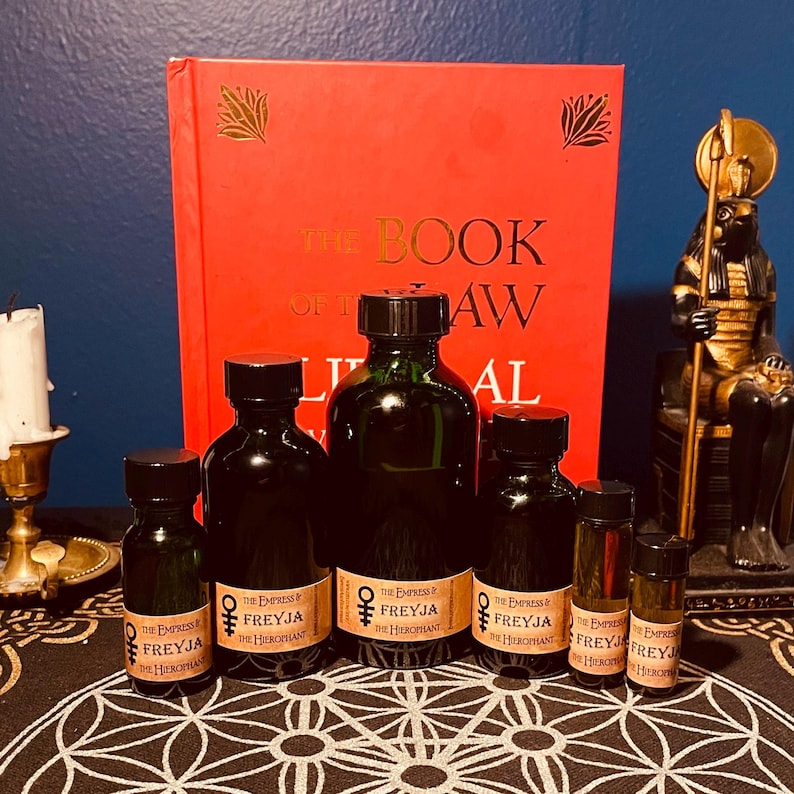 May include: A collection of six small glass bottles with brown labels. The labels feature the text "The Empress of Freyja The Hierophant". The bottles are arranged on a black and white patterned surface. A red book with gold lettering that reads "The Book of the Law" is visible in the background.