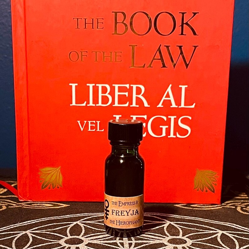 May include: A small dark brown glass bottle with a black cap and a label that reads "The Empress & Freyja The Hierophant". The bottle is sitting on a black and white patterned surface in front of a red book with the title "The Book of the Law Liber Al vel Legis".