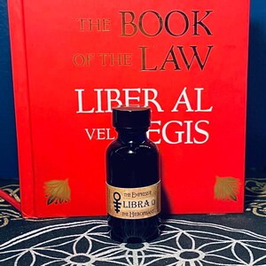 Puede incluir: Una peque&ntilde;a botella de vidrio oscuro con una tapa negra y una etiqueta que dice "THE EMPRESS & LIBRA THE HIEROPHANT". La botella est&aacute; delante de un libro rojo con el t&iacute;tulo "THE BOOK OF THE LAW LIBER AL VEL EGIS".