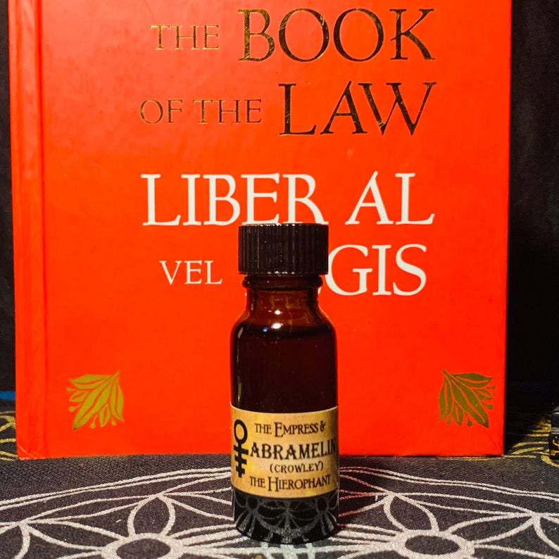 Puede incluir: Una peque&ntilde;a botella de vidrio marr&oacute;n con una tapa negra y una etiqueta que dice "THE EMPRESS & ABRAMELIN (CROWLEY) THE HIEROPHANT". La botella est&aacute; delante de un libro rojo con el t&iacute;tulo "THE BOOK OF THE LAW LIBER AL VEL GIS".