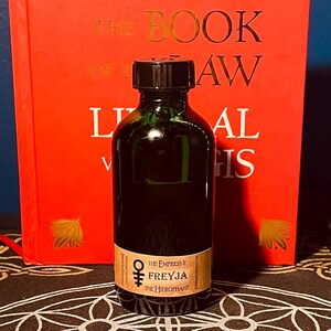 May include: A dark green glass bottle with a black cap. The bottle has a label that reads "The Empress & Freyja The Hierophant".