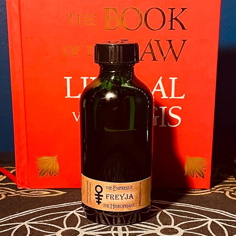 May include: A dark green glass bottle with a black cap. The bottle has a label that reads "The Empress & Freyja The Hierophant".