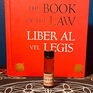 May include: A small glass bottle with a black cap and a brown liquid inside. The bottle is labeled "The Empress & Freyja Hierophant". The bottle is sitting on a black and white patterned surface in front of a red book titled "The Book of the Law Liber Al vel Legis".