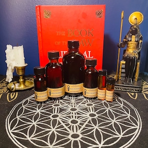 May include: An arrangement of dark glass bottles with black caps, labeled "The Empress & Abundance the Hierophant", are displayed in front of a red book titled "The Book of the Law". A candle in a brass holder and a statue of a seated deity are also visible.