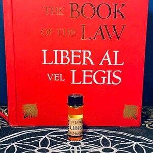 Puede incluir: Una peque&ntilde;a botella de vidrio transparente con una tapa negra llena de un l&iacute;quido dorado. La botella est&aacute; etiquetada con "The Empire of Libra, The Hierophant". La botella est&aacute; sobre una superficie con un patr&oacute;n blanco y negro delante de un libro rojo titulado "The Book of the Law, Liber Al vel Legis".