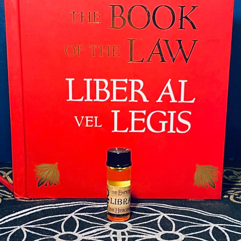 Puede incluir: Una peque&ntilde;a botella de vidrio transparente con una tapa negra llena de un l&iacute;quido dorado. La botella est&aacute; etiquetada con "The Empire of Libra, The Hierophant". La botella est&aacute; sobre una superficie con un patr&oacute;n blanco y negro delante de un libro rojo titulado "The Book of the Law, Liber Al vel Legis".