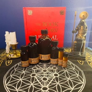 May include: An assortment of dark glass bottles with black caps, labeled "The Empress Abramelin" and a red book titled "The Book of the Law" are arranged on a black cloth with a geometric pattern. A candle and a statue are also present.