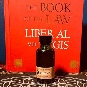 May include: A small dark brown glass bottle with a black cap. The bottle is labeled "The Empress & Freyja The Hierophant". The bottle is sitting in front of a red book with the title "The Book of the Law Liber Al vel Legis".