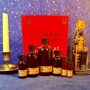 May include: An assortment of dark red glass bottles with black caps, labeled "The Empress & The Hierophant RAW POWER", are arranged in front of a red book titled "The Book of Law". A golden statue and a yellow candle are also in the image.