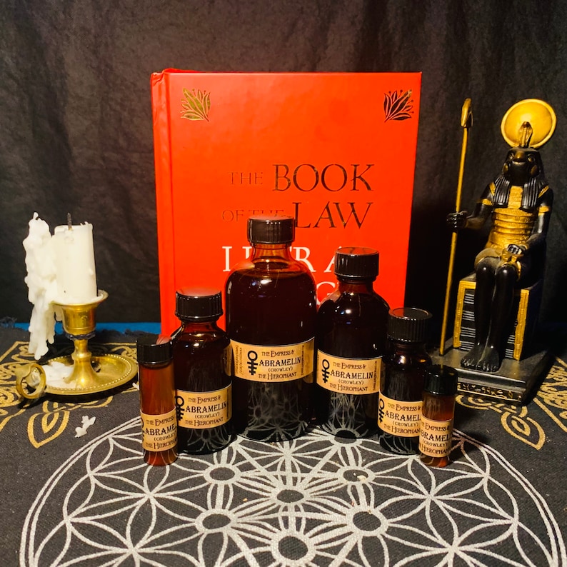 Puede incluir: Una colecci&oacute;n de seis botellas de vidrio &aacute;mbar con etiquetas que dicen "The Empress & Abramelin Crowley" y "The Hierophant". Las botellas est&aacute;n dispuestas sobre una superficie con estampado en blanco y negro delante de un libro rojo titulado "The Book of the Law" y una estatua dorada de una deidad egipcia.