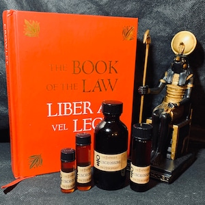 Könnte beinhalten: Vier kleine Glasflaschen mit brauner Flüssigkeit, jede mit dem Etikett "The Empress & Uncrossing The Hierophant". Die Flaschen stehen auf einem Tisch vor einem roten Buch mit dem Titel "The Book of the Law Liber AL vel Legis" und einer goldenen Statue eines ägyptischen Gottes.