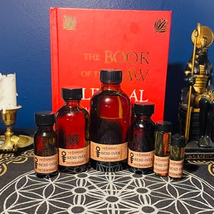 May include: Five glass bottles with dark red liquid and gold labels. The labels have the text "The Empress & The Hierophant" and "Bend Over". The bottles are arranged on a black and white patterned surface.
