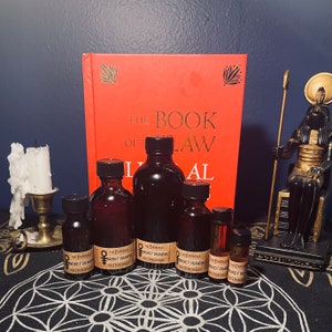 May include: A collection of six small glass bottles with brown liquid and labels that read "The Empress & The Hierophant Money Drawing" on a black and white patterned surface. The bottles are in front of a red book titled "The Book of Law" and a gold statue of an Egyptian god.