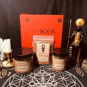 May include: Two glass jars of incense with black lids and labels that read "The Empress & The Hierophant Hadit Fine Handcrafted Incense". A red book with the title "The Book of" is visible in the background. A golden statue of an Egyptian god is also visible in the background.