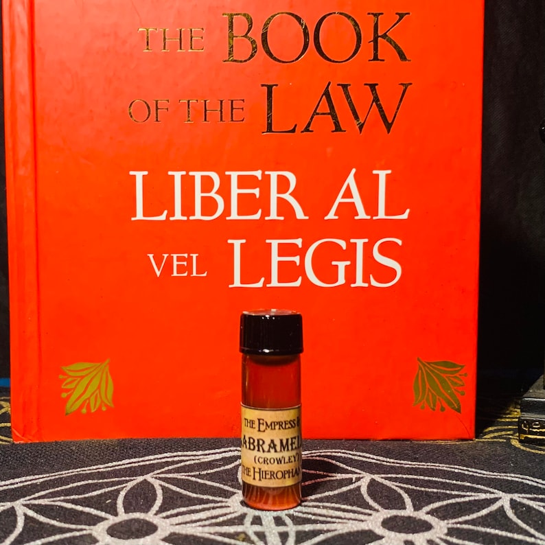 Puede incluir: Una peque&ntilde;a botella de vidrio transparente con una tapa marr&oacute;n oscura. La botella est&aacute; llena de un l&iacute;quido marr&oacute;n oscuro. La etiqueta de la botella dice "THE EMPRESS ABRAME (CROWLEY) THE HIEROPHA". La botella est&aacute; sobre una superficie con un patr&oacute;n blanco y negro delante de un libro rojo con el t&iacute;tulo "THE BOOK OF THE LAW LIBER AL VEL LEGIS".