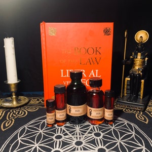 May include: Five small glass bottles with dark liquid inside. The bottles are labeled with the text "The Empress" and "Isis the Hierophant". A red book with gold lettering titled "The Book of the Law" is in the background. A black statue of an Egyptian god is on the right side of the image.
