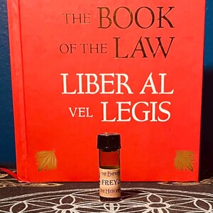 May include: A small, dark brown glass bottle with a black cap. The bottle is filled with a dark liquid. The bottle is labeled "The Empress Freyja The Hierophant". The bottle is sitting in front of a red book with the title "The Book of the Law Liber Al vel Legis".