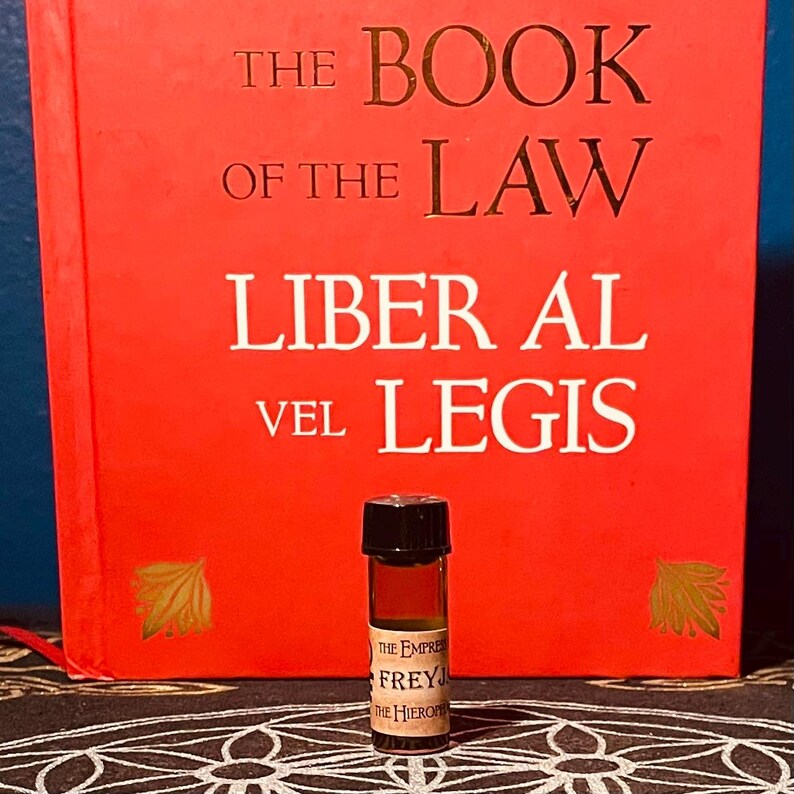 May include: A small, dark brown glass bottle with a black cap. The bottle is filled with a dark liquid. The bottle is labeled "The Empress Freyja The Hierophant". The bottle is sitting in front of a red book with the title "The Book of the Law Liber Al vel Legis".