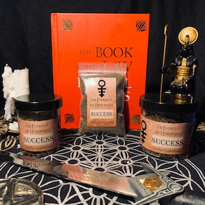 May include: Two small jars of incense labeled "The Empress & The Hierophant Success Fine Handcrafted Incense" are on a black and white patterned surface. A book titled "The Book of Law" is in the background.