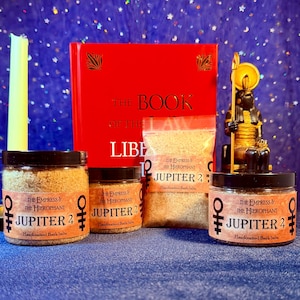 May include: Various jars and a bag of handcrafted bath salts, labelled "The Empress & The Hierophant Jupiter 2". A red book titled "The Book of the Law" is visible, alongside a golden statue and a yellow candle.