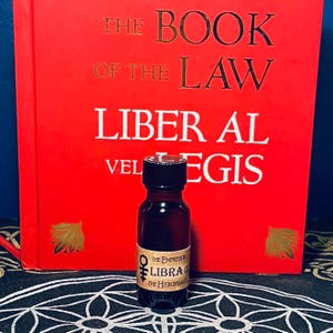 Puede incluir: Una peque&ntilde;a botella de vidrio marr&oacute;n con una tapa negra, etiquetada "The Empress & Libra 0 The Hierophant". La botella est&aacute; sentada sobre un patr&oacute;n circular blanco y negro, frente a un libro rojo titulado "The Book of the Law Liber Al vel Legis".