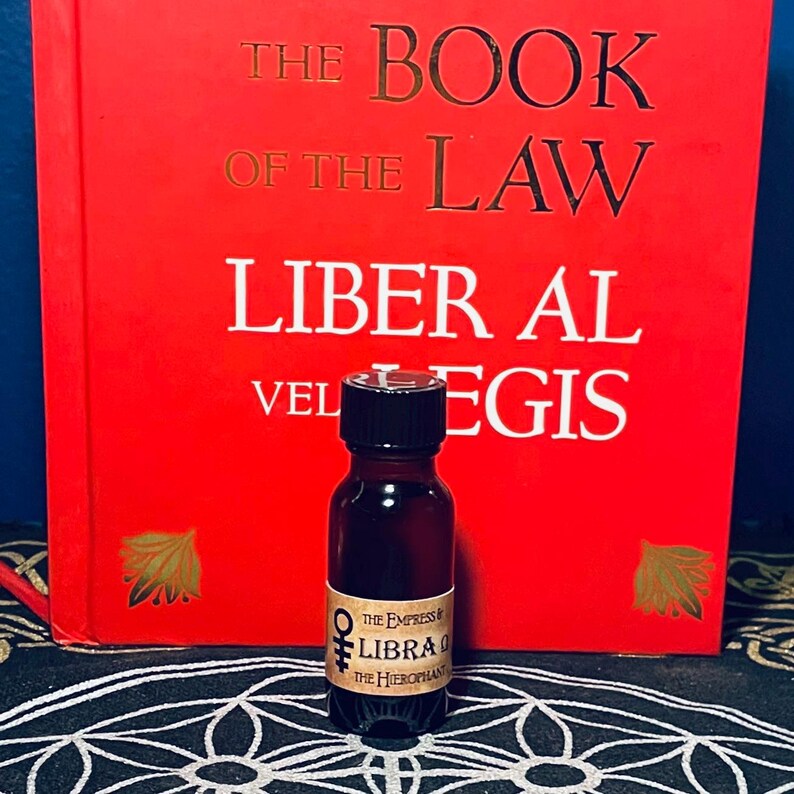 Puede incluir: Una peque&ntilde;a botella de vidrio marr&oacute;n con una tapa negra, etiquetada "The Empress & Libra 0 The Hierophant". La botella est&aacute; sentada sobre un patr&oacute;n circular blanco y negro, frente a un libro rojo titulado "The Book of the Law Liber Al vel Legis".