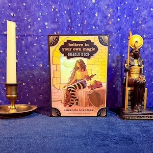 May include: A boxed oracle deck with the text "believe in your own magic" and an illustration of a woman with long hair. A candle and a statue of an Egyptian deity are on either side of the deck. The background is a blue fabric with white stars.