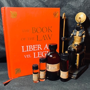 May include: Four small bottles of essential oil with labels that read "The Empress Road Opener The Hierophant". The bottles are arranged in a row on a dark surface. A red book with gold lettering that reads "The Book of the Law Liber Al vel Legis" is in the background. A gold statue of an Egyptian god is also in the background.
