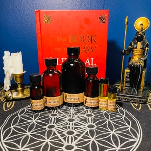May include: A collection of six amber glass bottles with labels that read "The Empress of Happiness by The Hierophant". The bottles are arranged on a black and white patterned surface with a red book and a gold statue of an Egyptian god in the background.