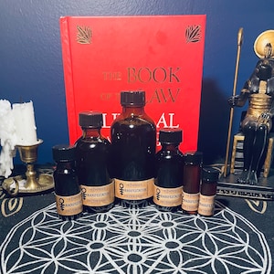 May include: Six small glass bottles with dark liquid inside, labeled "The Empress & The Hierophant Manifestation". The bottles are arranged on a black and white circular pattern with a red book titled "The Book of Law" in the background.