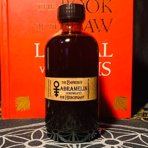 Puede incluir: Una botella de vidrio marr&oacute;n oscuro con una tapa negra y una etiqueta que dice "The Empress & Abramelin (Crowley) The Hierophant". La botella est&aacute; sobre una superficie con un patr&oacute;n blanco y negro.