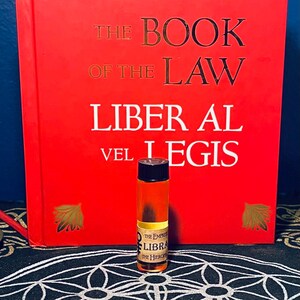 Puede incluir: Una peque&ntilde;a botella de vidrio &aacute;mbar con una tapa negra y una etiqueta que dice "The Empress Libra The Hierophant". La botella est&aacute; delante de un libro rojo con el t&iacute;tulo "The Book of the Law Liber Al vel Legis".
