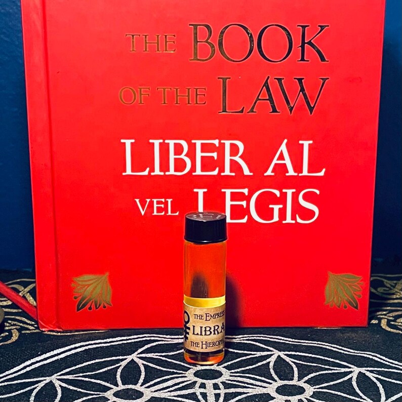 Puede incluir: Una peque&ntilde;a botella de vidrio &aacute;mbar con una tapa negra y una etiqueta que dice "The Empress Libra The Hierophant". La botella est&aacute; delante de un libro rojo con el t&iacute;tulo "The Book of the Law Liber Al vel Legis".