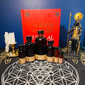May include: Six small brown glass bottles with labels that read "The Empress & Success The Hierophant" on a black and white patterned surface. A red book with the text "The Book of Law" is in the background.