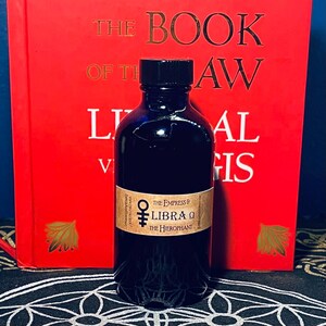 Puede incluir: Una botella de vidrio oscuro con una tapa negra, etiquetada "The Empress & LIBRA &Omega; The Hierophant". La botella est&aacute; sobre una superficie con un patr&oacute;n blanco y negro.