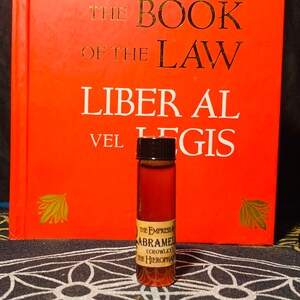 Puede incluir: Una peque&ntilde;a botella de vidrio marr&oacute;n con una tapa negra, que contiene un l&iacute;quido oscuro. La botella tiene una etiqueta con el texto "THE EMPRESS ABRAMEL (CROWLEY) THE HIEROPHAN". La botella est&aacute; sobre una superficie con un patr&oacute;n blanco y negro delante de un libro rojo con el t&iacute;tulo "THE BOOK OF THE LAW LIBER AL VEL LEGIS".