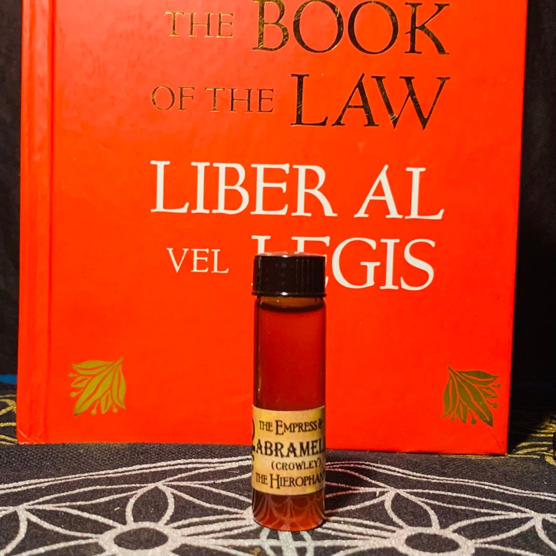 Puede incluir: Una peque&ntilde;a botella de vidrio marr&oacute;n con una tapa negra, que contiene un l&iacute;quido oscuro. La botella tiene una etiqueta con el texto "THE EMPRESS ABRAMEL (CROWLEY) THE HIEROPHAN". La botella est&aacute; sobre una superficie con un patr&oacute;n blanco y negro delante de un libro rojo con el t&iacute;tulo "THE BOOK OF THE LAW LIBER AL VEL LEGIS".