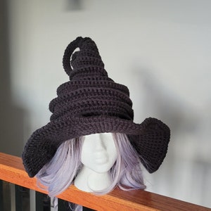 Puede incluir: Un sombrero de bruja de crochet negro con una corona alta y puntiaguda y un ala ancha. El sombrero se lleva en un maniqu&iacute; con cabello morado.