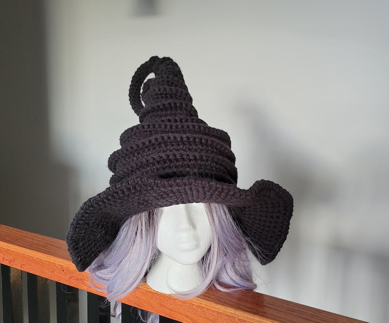 Puede incluir: Un sombrero de bruja de crochet negro con una corona alta y puntiaguda y un ala ancha. El sombrero se lleva en un maniqu&iacute; con cabello morado.