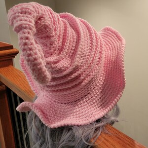 May include: A light pink, handmade crochet hat with a conical shape and a wide, ruffled brim. The hat is made of soft yarn and features a spiral design. The hat is shown on a person's head, resting on a wooden banister.