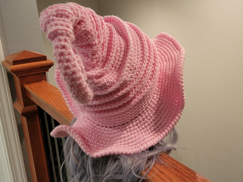 May include: A light pink, handmade crochet hat with a conical shape and a wide, ruffled brim. The hat is made of soft yarn and features a spiral design. The hat is shown on a person's head, resting on a wooden banister.