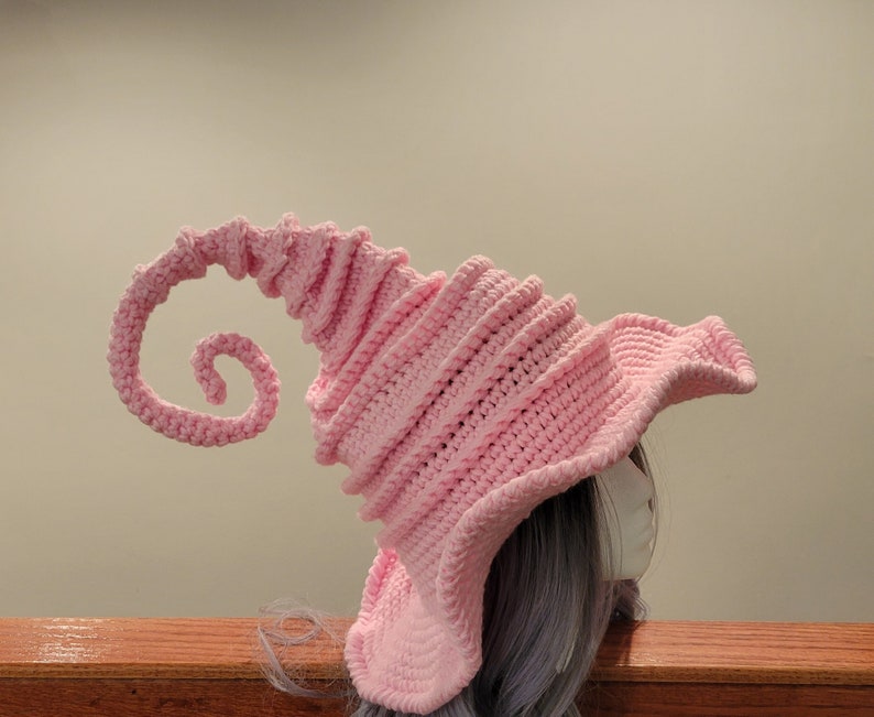 May include: A light pink, crocheted hat with a whimsical, swirled design. The hat features a wide brim and a long, spiraling extension. The soft, textured yarn creates a unique, fantasy-inspired accessory.
