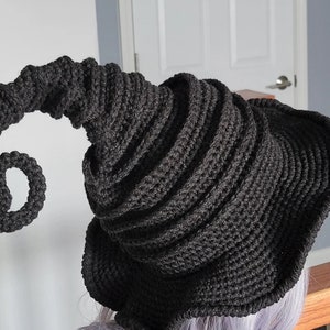 Puede incluir: Un sombrero de bruja de crochet negro con un ala larga y rizada. El sombrero est&aacute; hecho con un hilo grueso y tiene un dise&ntilde;o texturizado en espiral.
