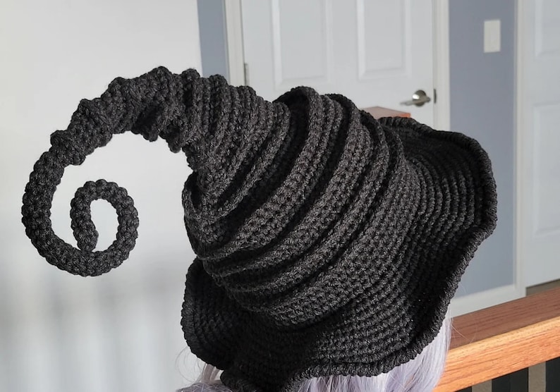 Puede incluir: Un sombrero de bruja de crochet negro con un ala larga y rizada. El sombrero est&aacute; hecho con un hilo grueso y tiene un dise&ntilde;o texturizado en espiral.