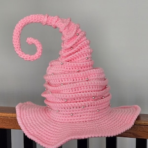 May include: A pink, crocheted witch hat with a curled tip and silver bead accents. The hat has a wide brim and a conical shape, perfect for Halloween or costume parties. The hat is made of soft yarn.