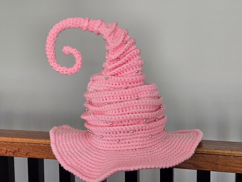 May include: A pink, crocheted witch hat with a curled tip and silver bead accents. The hat has a wide brim and a conical shape, perfect for Halloween or costume parties. The hat is made of soft yarn.