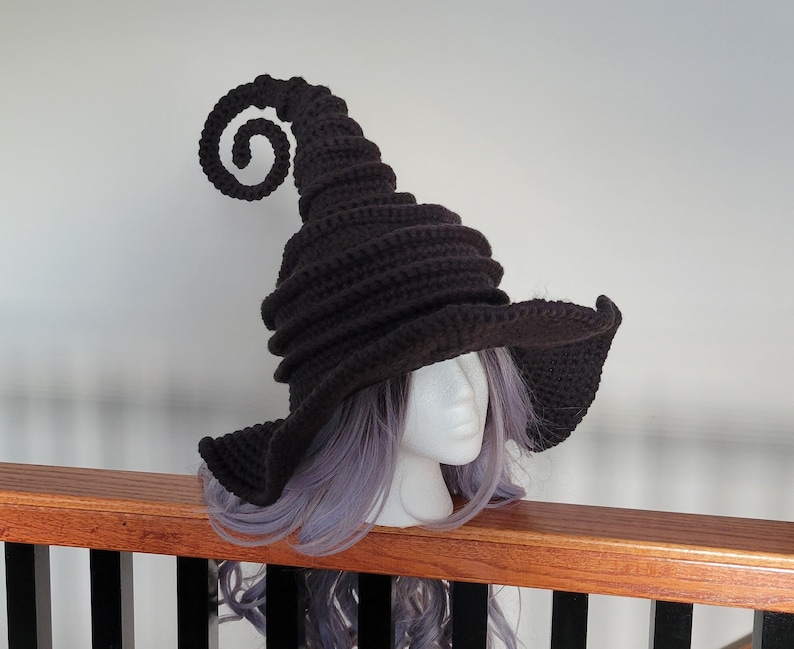 Puede incluir: Un sombrero de bruja de crochet negro con un borde rizado y una corona alta y puntiaguda. El sombrero se lleva en un maniqu&iacute; con cabello morado.