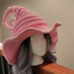 May include: A light pink, crocheted witch hat with a wide brim and a curled, horn-like detail. The hat is displayed on a mannequin head with gray hair. The hat's design is whimsical and suitable for costume or cosplay.