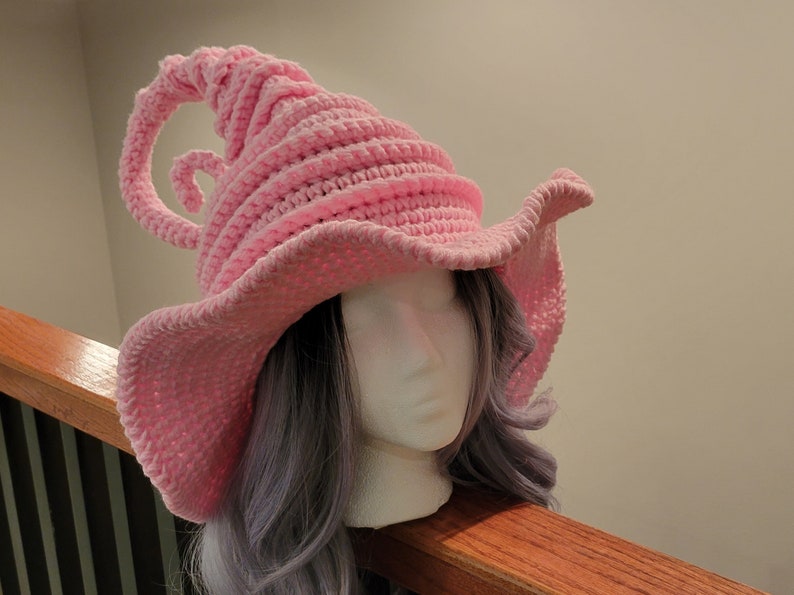 May include: A light pink, crocheted witch hat with a wide brim and a curled, horn-like detail. The hat is displayed on a mannequin head with gray hair. The hat's design is whimsical and suitable for costume or cosplay.