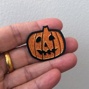 May include: A black and orange glitter pumpkin pin with a carved jack-o'-lantern face.